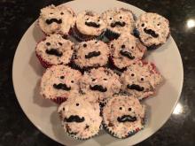 A plate containing a dozen cupcakes. The cupcakes are covered in fun-fetti icing, and decorated with candy eyeballs and moustaches to look like faces. Each cake's eyes and faces look sightly different, giving each one a unique expression, while appearing to look in different directions.